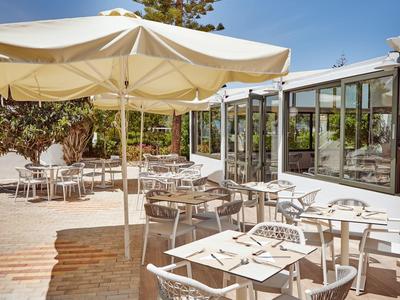 Freiluftcafé mit weißen Tischen, Stühlen und großen Sonnenschirmen bei klarem Himmel.