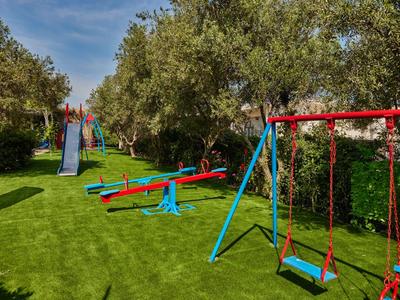 Spielplatz mit Schaukeln, Rutsche und Wippen auf grünem Rasen unter blauem Himmel und Bäumen.