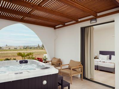 Terrasse mit Whirlpool, zwei Stühlen, offenem Schlafzimmer und Blick auf Landschaft unter blauem Himmel.