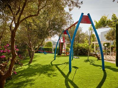 Kinderspielplatz mit Schaukeln auf grünem Rasen, umgeben von Bäumen und blühenden Sträuchern.
