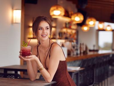 Eine lächelnde Frau in elegantem Kleid sitzt an einer Bar und hält ein Getränk mit Minze.