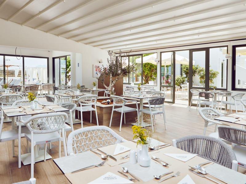 Restaurante moderno y luminoso con mesas blancas y grandes ventanas con vistas a la terraza y la ciudad.