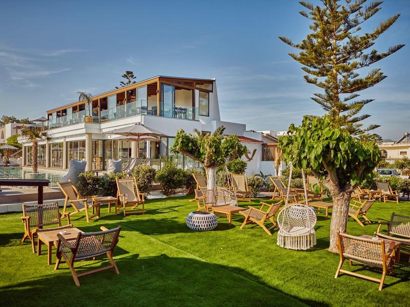 Hotel moderno con jardín, sillas de madera y hamacas bajo un cielo azul.