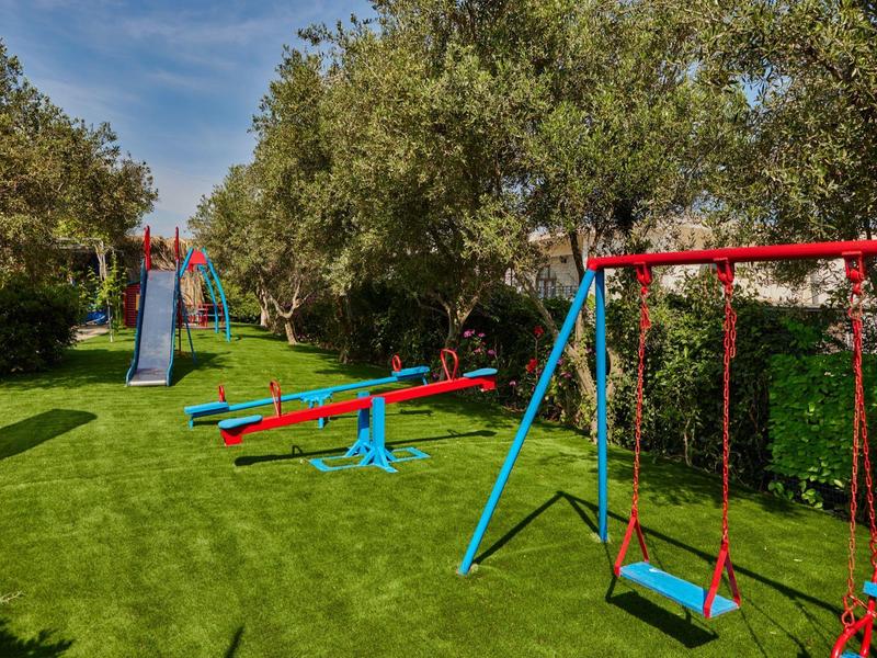 Área de juegos infantiles con columpios, balancines y estructura para trepar rodeada de árboles sobre césped verde.