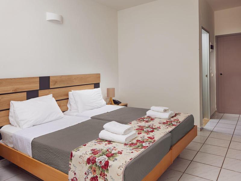 Una habitación de hotel simple con cama doble, ropa blanca y cubrecolchón gris, piso de baldosas.