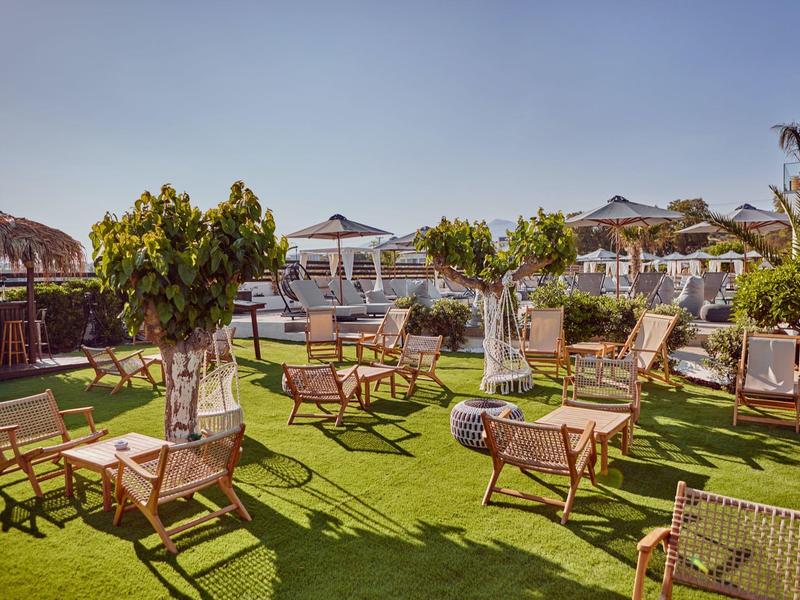 Área de asientos al aire libre con sillas y mesas de madera sobre césped verde bajo sombrillas.