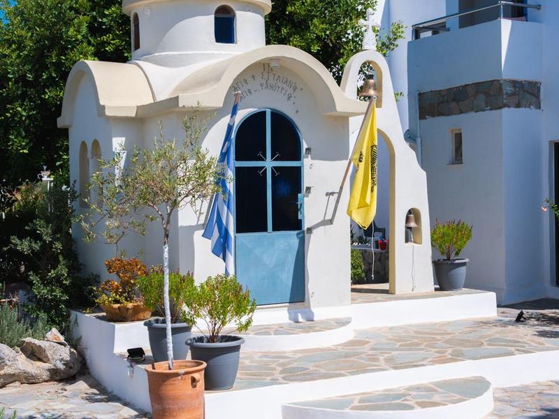 Pequeña iglesia blanca con puerta azul y campanario frente a edificio blanco bajo cielo despejado.