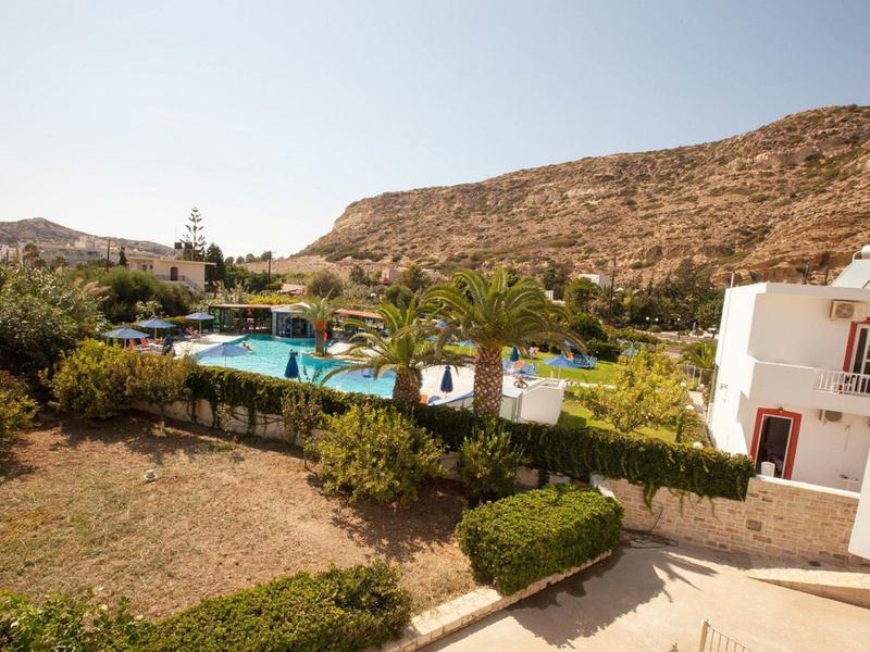 Hotel met zwembad, omgeven door groene struiken en bergen op de achtergrond.
