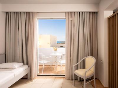 Lichte hotelkamer met eenpersoonsbed, stoel en balkon met uitzicht op gebouwen en zee.