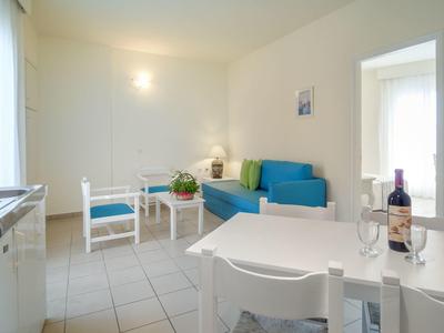 Helles Wohnzimmer mit blauer Couch, zwei weißen Stühlen und Esstisch mit Weinflasche.