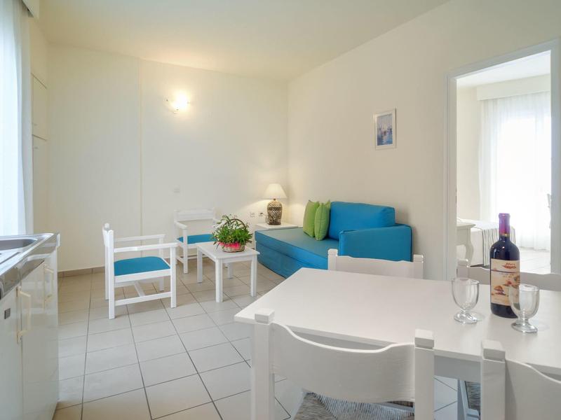 Helles Wohnzimmer mit blauer Couch, zwei weißen Stühlen und Esstisch mit Weinflasche.