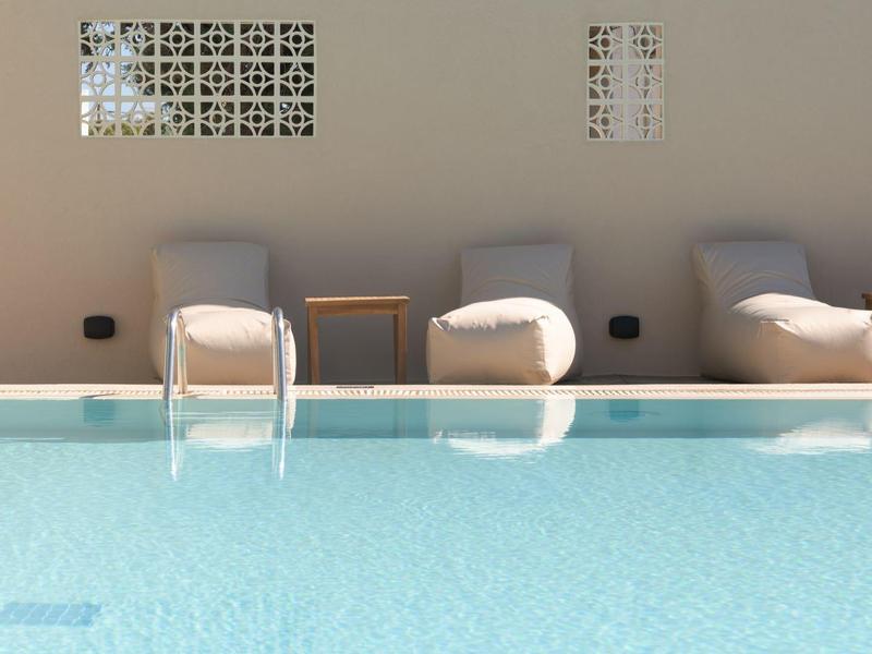 Niebieski basen z dwoma drabinkami przed ścianą z trzema leżakami i ozdobnymi wentylacjami.