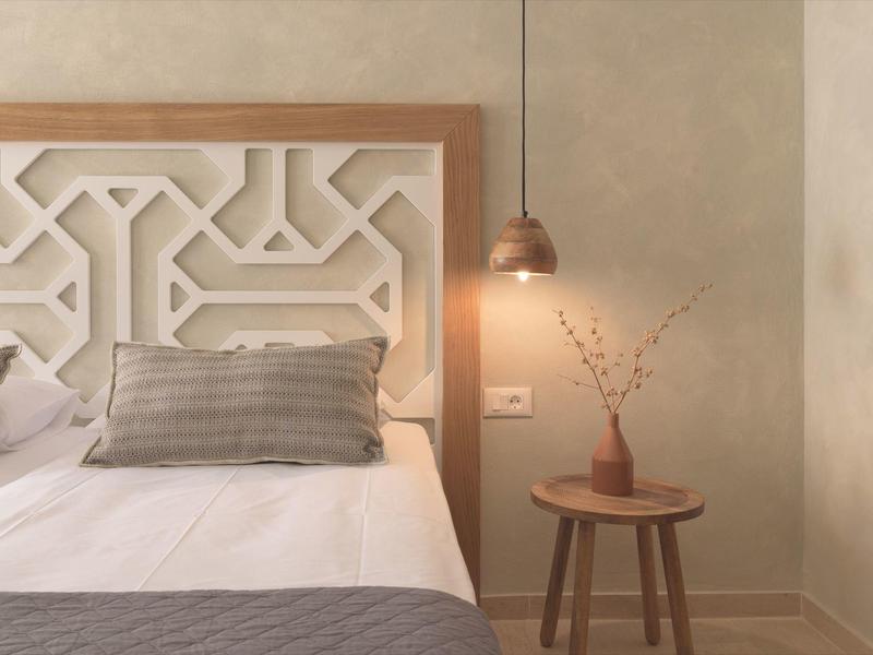 Przytulna sypialnia z białym łóżkiem, szarymi poduszkami i małym drewnianym stolikiem z wazonem.