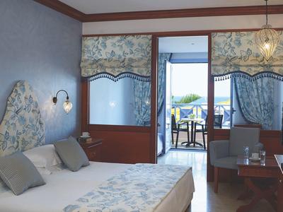 Bright hotel room with double bed, seating area, and balcony overlooking the sea.