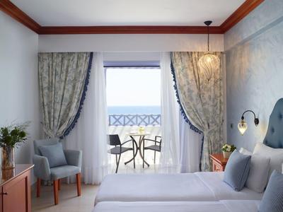 Bright hotel room with double bed, balcony, and seating area overlooking the sea.