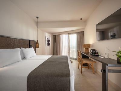 Modernes Hotelzimmer mit großem Bett, Schreibtisch, Fernseher und Balkon mit Vorhängen.