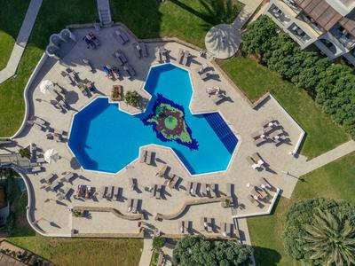 Poolbereich mit blauem Wasser, umgeben von Liegestühlen auf hellem Boden, umgeben von Grünflächen.