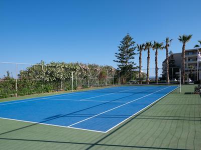 Ein blauer Tennisplatz mit grünem Umrandung und Palmen im Hintergrund neben einem Hotel.