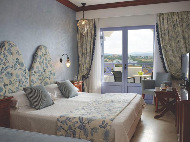 Brightly furnished hotel room with double bed, balcony, and view of the landscape.