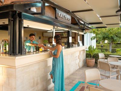 Een vrouw in een blauwe jurk staat bij een buitensbar met een barman en tafels op de achtergrond.