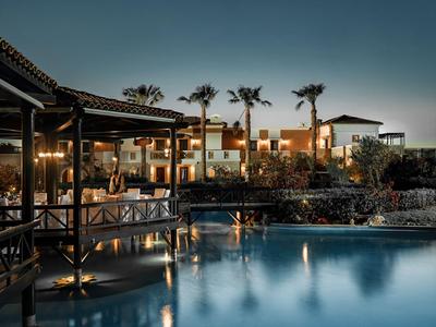 Abendansicht eines beleuchteten Hotels mit Pool und Palmen unter klarem Himmel.