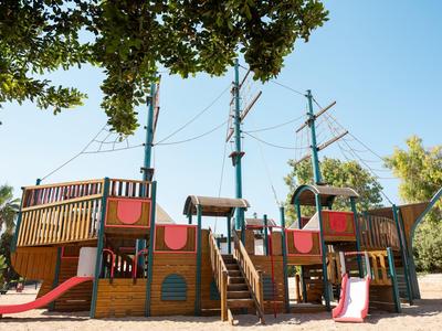 Kinderspeelplaats in de vorm van een groot houten schip met glijbanen en klimtoestellen onder bomen.