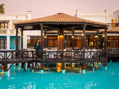 Een hotelrestaurant op palen boven een turquoise zwembad bij zonsondergang.