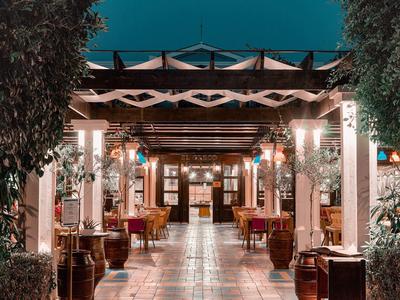 Osvětlený otevřený průchod se stoly a židlemi, lemovaný rostlinami, vedoucí k budově.