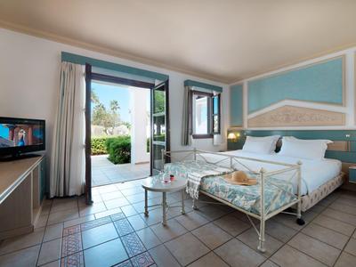 Lichte hotelkamer met wit metalen bed, tegelvloer en open deur naar tuin met groen.