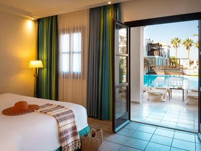 Licht ingerichte hotelkamer met uitzicht op zwembad en palmbomen door een open deur.