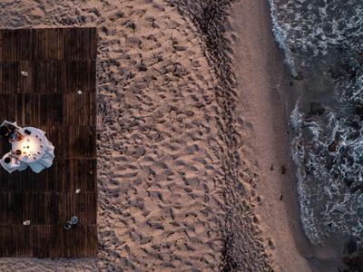 Houten terras op zanderig strand naast golven, persoon in witte jurk zittend op vloer.