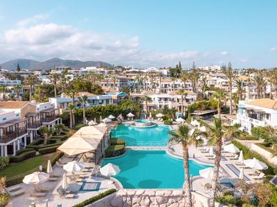 Rezort s velkým bazénem, lehátky, palmami a bílými budovami pod modrou oblohou.