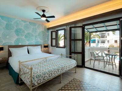 Světle hotelový pokoj s vzorovaným přehozem a otevřenými dveřmi na terasu s stolem a židlemi.