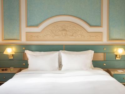 Elegantní hotelová postel s bílým povlečením, dvěma polštáři, dřevěným čelem a zabudovanými lampami.