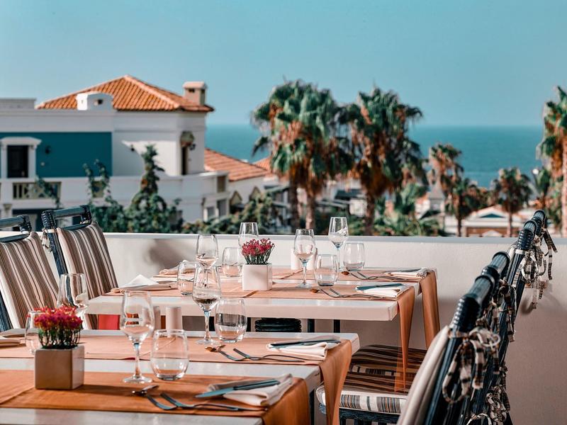 Venkovní jídelna s prostřenými stoly, židlemi, palmami a výhledem na oceán.