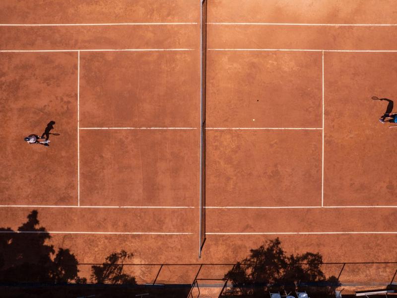 Ptačí pohled na dvě osoby hrající tenis na přilehlých antukových kurtech se stíny a viditelnou sítí.