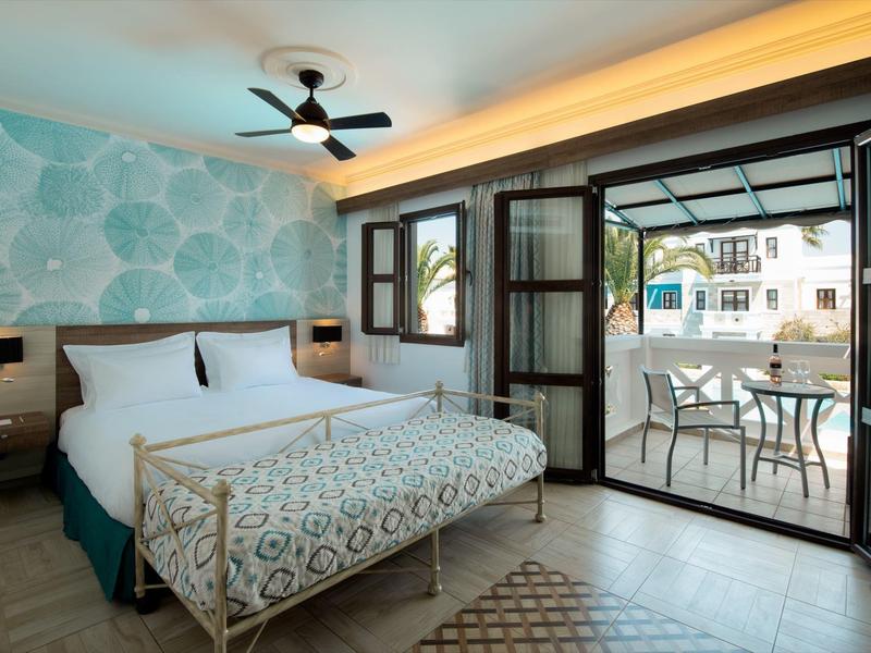 Světle hotelový pokoj s vzorovaným přehozem a otevřenými dveřmi na terasu s stolem a židlemi.