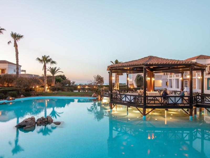 Bazénová oblast resortu s palmami, lehátky a altánem nad klidnou modrou vodou za soumraku.