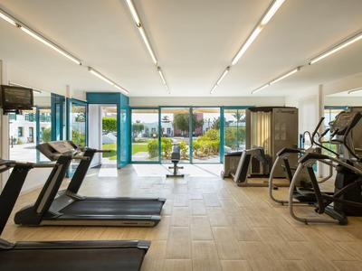 Heller Fitnessraum mit Laufbändern, Crosstrainern und Panoramafenstern mit Blick auf eine Grünfläche.