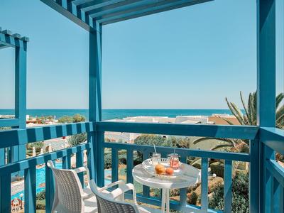 Balkon mit blauem Geländer, kleinem Tisch mit Getränken, Blick auf Meer und Häuser bei klarem Himmel.