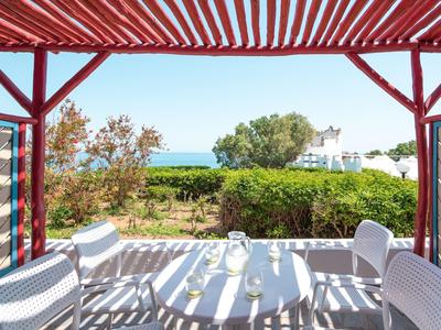 Terrasse mit weißem Tisch und Stühlen unter rotem Sonnenschutz, Blick auf grüne Pflanzen und Meer.
