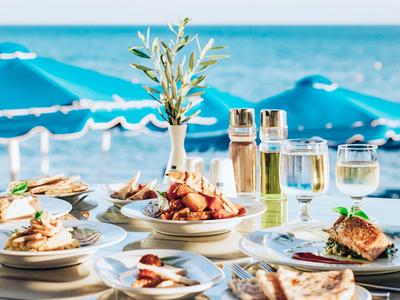 Tisch mit kulinarischen Gerichten und Getränken am Strand vor blauen Sonnenschirmen.