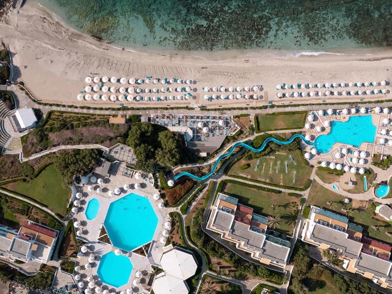 Luftaufnahme eines Resorts mit mehreren Pools, Liegestühlen am Strand und klarem türkisfarbenem Wasser.