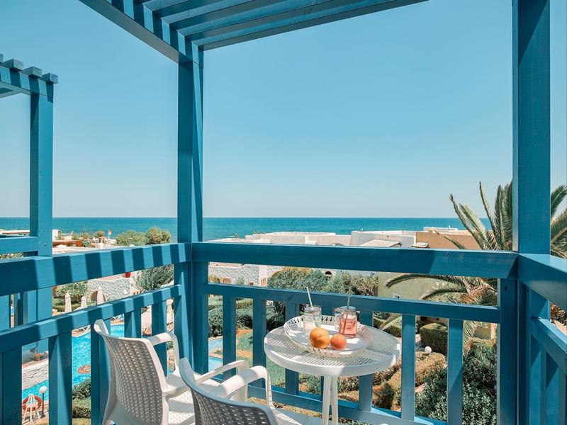 Balkon mit blauem Geländer, kleinem Tisch mit Getränken, Blick auf Meer und Häuser bei klarem Himmel.