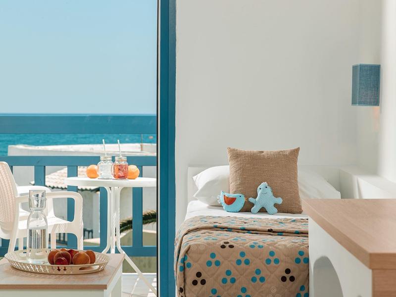 Balkon mit Meerblick, weißer Stuhl und Tisch mit Obst, innen Bett mit Kissen und blauem Stern.