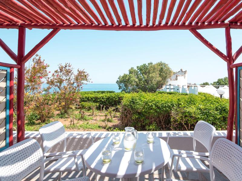 Terrasse mit weißem Tisch und Stühlen unter rotem Sonnenschutz, Blick auf grüne Pflanzen und Meer.