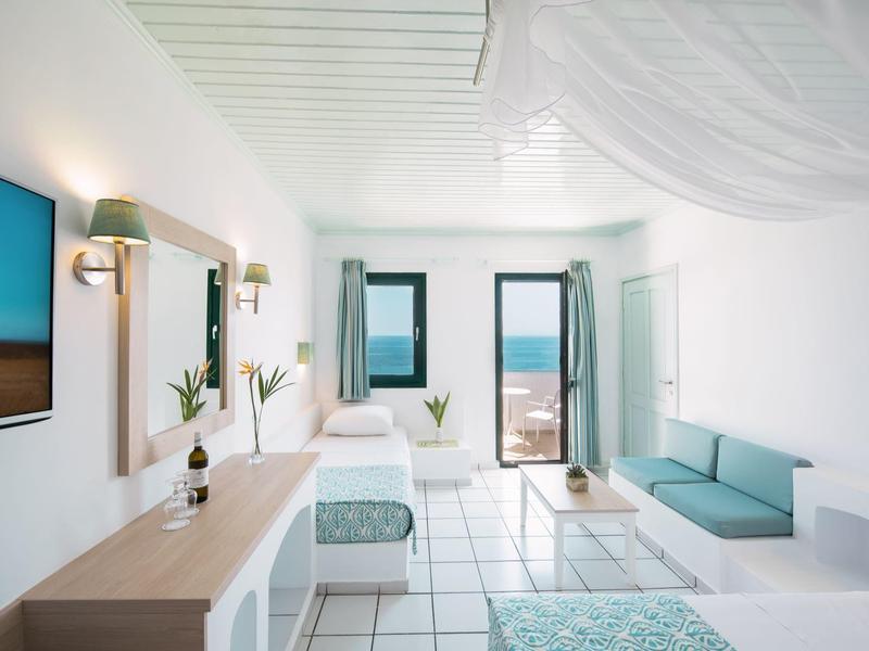 Helles, modernes Zimmer mit weißen Wänden, Bett, Sofa, Fernseher und Blick auf Meer durch Fenster.