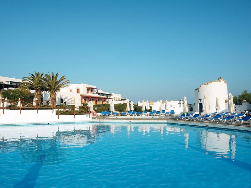 Großer blauer Swimmingpool mit weißen Häusern, Palmen und Sonnenschirmen unter klarem Himmel.