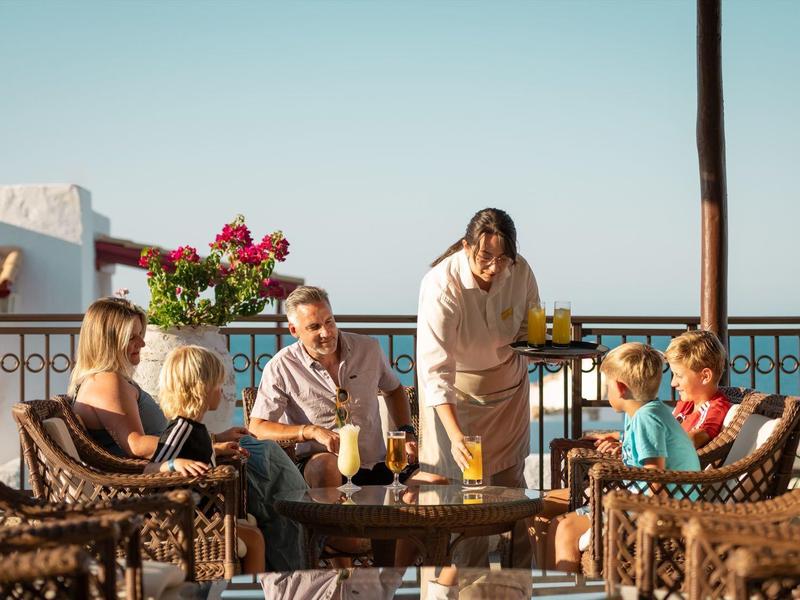 Eine Familie sitzt auf einem Balkon mit Meerblick, während ein Kellner ihnen Getränke serviert.