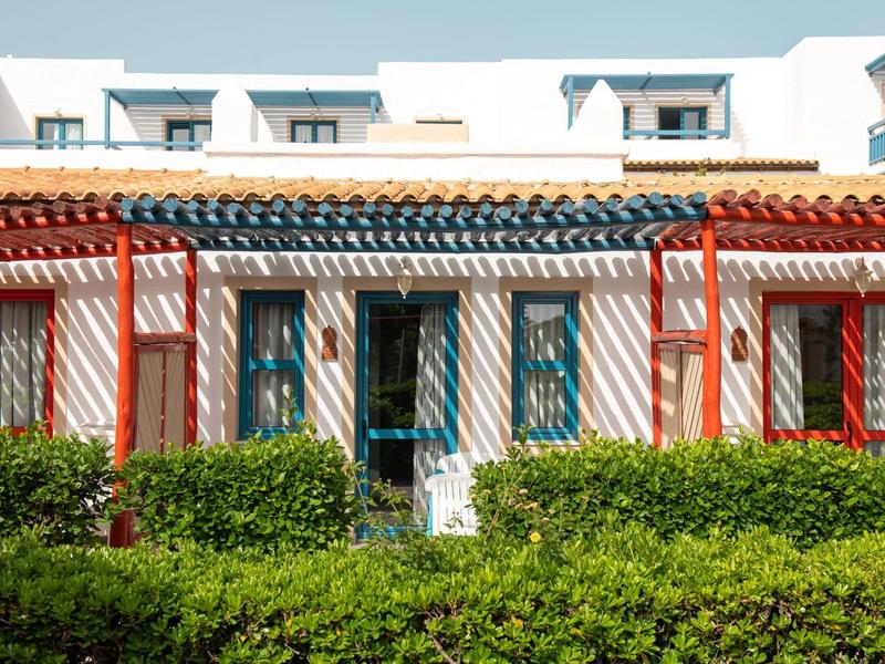 Haus mit weißer Fassade, blauen Türen und Fenstern, roten Jalousien und grünen Büschen davor.